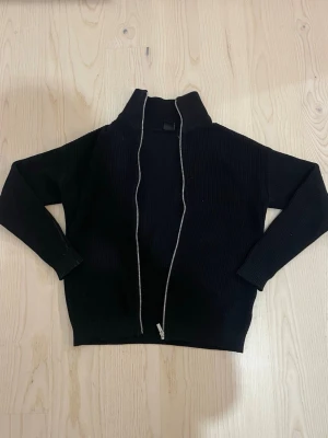 Svart ribbad ziptröja med hög krage - Säljer en svart ribbad ziptröja med hög krage och hel dragkedja framtill. Tröjan har långa ärmar och en enkel, stilren design som passar perfekt till lager på lager. Materialet känns mjukt och stretchigt, vilket gör den bekväm att bära.