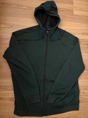 Mörkgrön hoodie från BOSS - Snygg mörkgrön hoodie från BOSS med dragkedja framtill och broderad logga på bröstet. Hoodien har huva med dragsko och ribbade muddar vid ärmslut och nederkant. Perfekt för en avslappnad och stilren look.