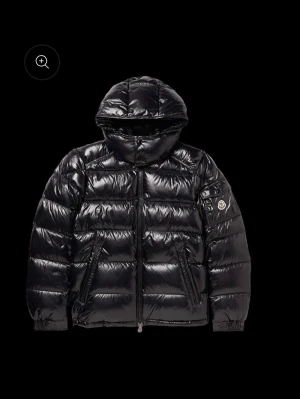 Svart dunjacka från Moncler - Snygg svart dunjacka från Moncler med glansig finish och huva. Jackan har två dragkedjefickor framtill, ribbade muddar och Moncler-logga på ärmen. Perfekt för kalla vinterdagar och riktigt trendig stil.
