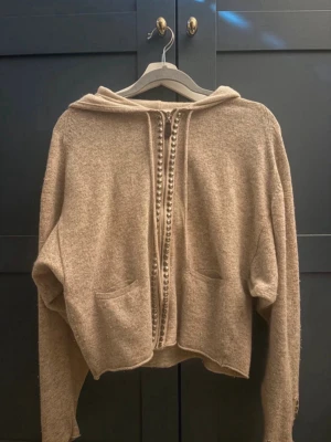 Beige kofta med huva från Hunkydory - Mysig beige kofta från Hunkydory med huva och dekorativa knappar längs hela framsidan. Koftan har två fickor framtill och är tillverkad i ett mjukt, stickat material. Har dessvärre två små hål på höger arms slut. 