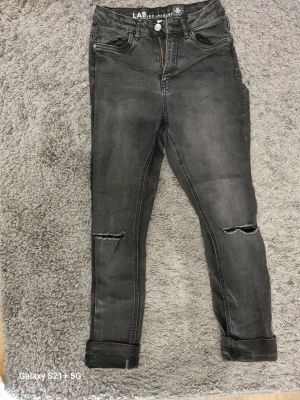 Grå slitna skinny jeans med hål - Säljer ett par grå skinny jeans med slitningar och hål på knäna. Jeansen har hög midja, fem fickor och uppvikta benslut. Perfekta för en avslappnad och trendig look. Materialet är stretchigt denim för extra komfort.