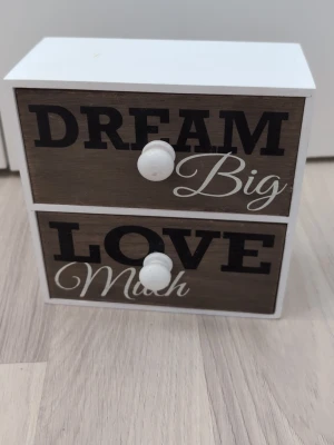 Liten låda med text dream love - Säljer en liten förvaringslåda i trä från ARTI CASA med två lådor. Framsidan har texten 'DREAM Big' och 'LOVE Much' i svart och vitt. Lådan är vit med träfärgade fronter och vita knoppar. Perfekt för småprylar eller smycken.