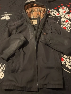 Äkta burberry vindjacka - Snygg svart vindjacka med dragkedja och huva. Insidan har ett klassiskt beige-rutigt foder som ger extra stilpoäng. Jackan har fickor framtill och justerbara ärmslut med knapp. Perfekt för blåsiga dagar och enkel att matcha med olika outfits. Den enda skada är den som syns på tredje bilden men det man märker knapp om man inte kolla nogrant