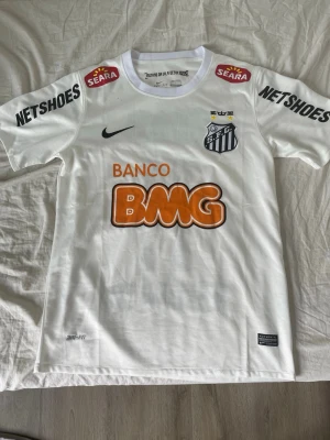 Vit Santos Neymar Jr matchtröja Nike - Säljer en vit Santos matchtröja från Nike med Neymar Jr och nummer 11 på ryggen. Tröjan har flera sponsorloggor i svart, orange och rött samt klubbmärke på bröstet. Tillverkad i lätt och ventilerande material, perfekt för fotboll eller samling.