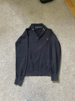 Mörkblå stickad tröja från GANT - Snygg mörkblå stickad tröja från GANT med halv zip och klassisk krage. Tröjan har diskret GANT-logga broderad på bröstet och ribbade muddar vid ärmslut och nederkant. Defekterna är att dragläppen är borta på dragkedjan men går fortfarande att använda och en liten fläck på sidan av magen. Perfekt för en stilren och avslappnad look.