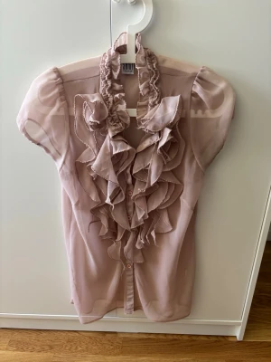 Ljusrosa volangblus i transparent tyg - Ljusrosa blus i transparent, lätt material med korta ärmar. Framtill har blusen stora volanger längs knäppningen och en rynkad hög krage. Perfekt för dig som gillar romantiska detaljer och en feminin look.