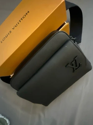 Svart Louis Vuitton crossbody-väska - Snygg svart crossbody-väska från Louis Vuitton i grained läder med präglad LV-logga framtill. Väskan har justerbar axelrem och svarta metalldetaljer med Louis Vuitton-gravyr. Perfekt för att bära det viktigaste med stil.