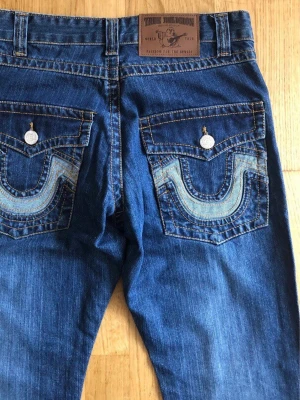 Blå True Religion jeans med kontrastsömmar - Säljer ett par klassiska blå jeans från True Religion med raka ben och tydliga kontrastsömmar. Bakfickorna har den ikoniska hästsko-designen och stora knappar. Storlek 28 tum. Mått: Ytterbenslängd 104 cm, Innerbenslängd 79 cm