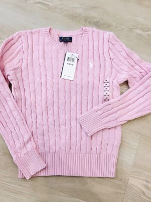 Ljusrosa kabelstickad tröja Polo Ralph Lauren - Ljusrosa kabelstickad tröja från Polo Ralph Lauren med rund halsringning och ribbade muddar vid ärmslut och nederkant. Klassisk broderad logga på bröstet. Perfekt för dig som gillar en stilren och tidlös look. Olika storlek finns 