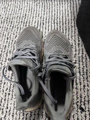 Grå Adidas Ultraboost sneakers - Säljer ett par grå Adidas Ultraboost sneakers med stickad ovandel och klassiska tre ränder på sidan. Skorna har grå snörning och en bekväm, stötdämpande sula. Perfekta för dig som gillar sportig och stilren look.