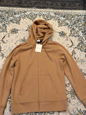 Beige hoodie med dragkedja från GANT med tag kvar  - Helt ny Snygg beige hoodie från GANT med dragkedja framtill och två fickor. Klassisk huva med dragsko och diskret GANT-logga på bröstet. Tillverkad i mjukt material som känns skönt mot huden. Perfekt för en avslappnad och stilren look.