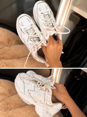 Vita New Balance sneakers med rosa detaljer - Säljer ett par vita New Balance sneakers med rosa detaljer och klassisk N-logga på sidan. Skorna har chunky sula, vita skosnören och är tillverkade i en mix av läder och syntetmaterial. Perfekta för en sportig och trendig look.