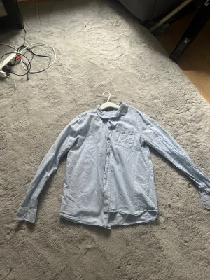 Ljusblå klassisk skjorta - OBS den är inte struken!! Säljer en ljusblå skjorta i bomull med klassisk krage och bröstficka. Skjortan har långa ärmar och knäppning framtill. Perfekt till jeans eller chinos för en clean och enkel stil.