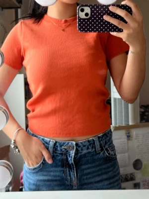 Orange ribbad croppad topp - Säljer en orange ribbad croppad topp med korta ärmar och rund hals. Toppen har en enkel och clean look som passar perfekt till jeans eller kjol. Materialet känns mjukt och stretchigt, vilket gör den bekväm att bära hela dagen.