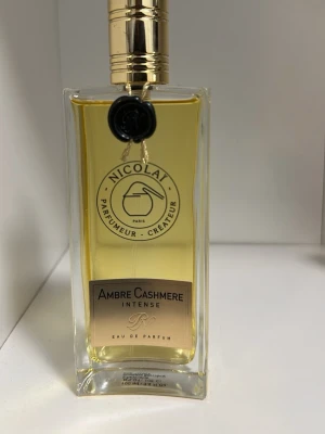 Ambre Cashmere Intense Eau de Parfum - Ambre Cashmere Intense Eau de Parfum från Nicolai i en elegant, rektangulär glasflaska med guldfärgat lock och detaljer. Doften har en varm, bärnstensgul vätska och flaskan pryds av en svart sigill och etikett framtill.