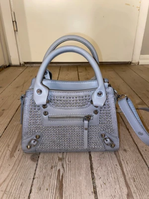 Ljusgrå handväska med nitar - Trendig ljusgrå handväska i konstläder med silverfärgade nitar på framsidan. Väskan har både handtag och justerbar axelrem. Perfekt för dig som gillar detaljer och vill sticka ut med din look.