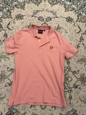 Rosa pikétröja från lyle and scott  - Snygg rosa pikétröja med klassisk krage och korta ärmar. Tröjan har en broderad gul logga på bröstet och knäppning framtill. Perfekt för en avslappnad dag