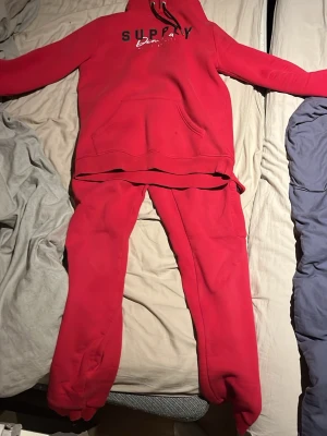 Rött loungewear-set med hoodie - Mysigt rött loungewear-set med matchande hoodie och byxor. Hoodien har magficka, dragsko i huvan och tryckt text på bröstet. Byxorna har resår i midjan och benslut för extra komfort. Perfekt för chill hemma eller slappa dagar.