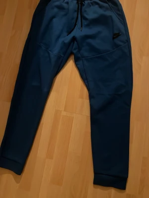 Blåa mjukisbyxor från Nike - Säljer ett par blåa mjukisbyxor från Nike med svart logga på vänster ben. Byxorna har dragsko i midjan, två sidofickor och muddar vid bensluten. Perfekta för chill eller träning, riktigt sköna och stilrena.