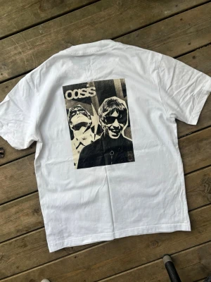 Vit Oasis t-shirt med tryck - Säljer en vit t-shirt med stort svartvitt tryck på ryggen föreställande bandet Oasis. T-shirten har klassisk passform och rund hals. Perfekt för dig som gillar bandmerch och vill ha en stilren men ändå uttrycksfull look.