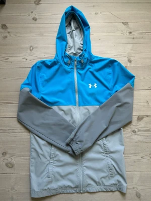 Blå och grå vindjacka från Under Armour - Snygg vindjacka från Under Armour i blått och grått. Jackan har huva med dragsko, hel dragkedja framtill och två sidofickor. Tillverkad i lätt och vindtätt material, perfekt för blåsiga dagar. Under Armour-logga på bröstet.