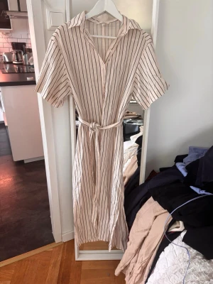 Randig långklänning med knytband - Säljer en beige långklänning med svarta vertikala ränder. Klänningen har korta ärmar, krage, knappar framtill och ett matchande knytband i midjan. Perfekt för en avslappnad men ändå stilsäker look. Linen blend.