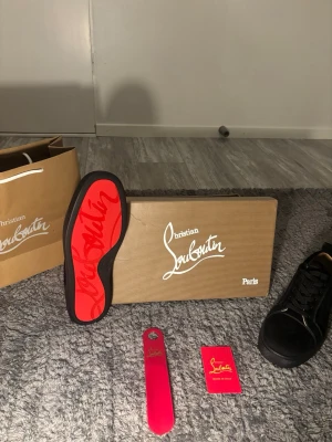  Christian Louboutin - Snygga svarta sneakers från Christian Louboutin med klassisk röd sula och svarta skosnören. Skorna har en stilren design i mocka och läder, samt diskreta detaljer. Perfekta för dig som vill ha något exklusivt och trendigt i garderoben.