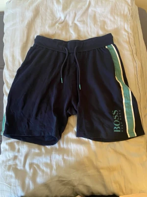 Svarta shorts från Hugo Boss - Snygga svarta shorts från Hugo Boss med gröna och vita ränder på sidorna samt logga på vänster ben. Shortsen har snörning i midjan och är perfekta för en avslappnad stil. Tillverkade i mjuk bomull för extra komfort.