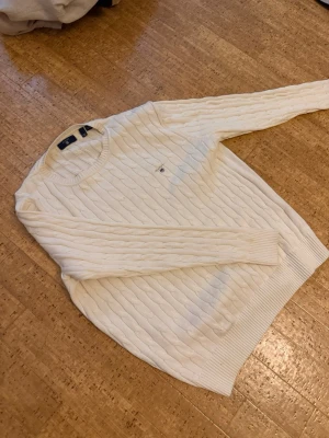 Kabbelstickad Gant tröja - Kabelstickad vit tröja med rund halsringning och ribbade muddar. Tröjan har ett diskret broderat emblem på bröstet och är tillverkad i ett mjukt material som passar perfekt för kyligare dagar. Klassisk och stilren design med lång ärm. Billigare vid snabba köp.
