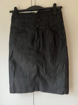Mörkblå jeanskjol med knappar - Snygg mörkblå jeanskjol med hög midja i rak modell med tre knappar framtill, fickor både fram och bak. Kjolen har en slits bak
