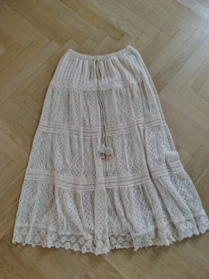Beige maxikjol med spets från Chica London - Romantisk beige långkjol med vacker spets och volangkant. Kjolen har knytband i midjan och ett detaljerat spetsmönster som ger en bohemisk vibe. Perfekt för dig som gillar en feminin och avslappnad stil.