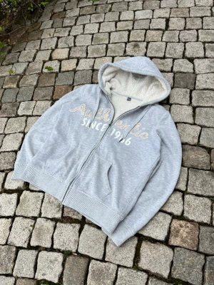Ljusgrå hoodie från Pimkie - Ljusgrå hoodie med dragkedja och broderad text 'Authentic Since 1966' på bröstet. Mjuk teddyfoder i huvan och två fickor framtill. Ribbstickade muddar vid ärmslut och nederkant. Perfekt för chill dagar och streetwear-stil.