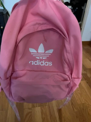 Rosa ryggsäck från Adidas - Snygg rosa ryggsäck från Adidas med det klassiska vita treklöver-loggan på framsidan. Ryggsäcken har ett stort huvudfack och en mindre ytterficka med dragkedja. Axelremmarna är justerbara och har Adidas-tryck. Perfekt för skola eller träning. Den har lite fläckar som inte går bort 