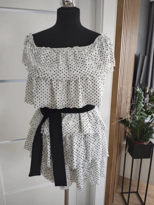  Ny slutsåld Prickig volangklänning med svart bälte - Söt kort offshoulder-klänning i vitt med svarta prickar och flera volanglager. Klänningen har ett brett svart knytbälte i midjan och är tillverkad i ett lätt, luftigt material. Perfekt för dig som gillar romantisk och trendig stil.