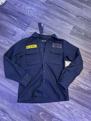 Svart overshirt från Replay - Snygg svart overshirt från Replay med två bröstfickor och coola patchar, bland annat 'HELL ON WHEELS'. Stora Replay-trycket på ryggen. Overshirten har knäppning framtill och är gjord i ett slitstarkt bomullstyg. Perfekt för en streetwear-look.