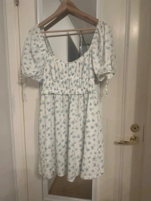 Blommig kortklänning från Gina Tricot - Supersöt vit kortklänning från Gina Tricot med blått blommönster. Klänningen har puffärmar med knytband, smockad midja och en luftig, flowy passform. Perfekt för varma dagar och somriga vibes! Endast använd en gång 