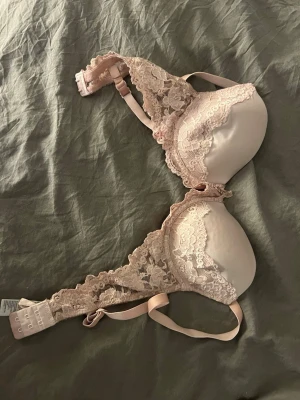 Ljusrosa spets-bh med vaddering - Söt ljusrosa bh med vadderade kupor och dekorativa spetsdetaljer längs kanterna och banden. Bh:n har justerbara axelband och hakknäppning bak. Materialet är en kombination av mjuk spets och slät textil för extra komfort och stil.