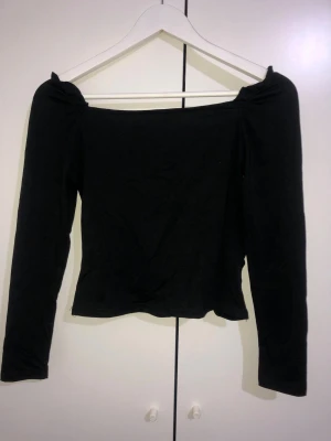 Svart off shoulder topp med rynk - Trendig svart off shoulder topp med långa ärmar och rynkade detaljer framtill. Toppen har en figurnära passform och är tillverkad i ett mjukt stretchigt material som sitter snyggt på kroppen. Perfekt för dig som gillar en stilren men ändå cool look.