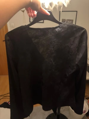 Svart blus med knyt framtill - Snygg svart blus från Gina Tricot i glansigt tyg med diskret blommönster. Blusen har djup v-ringning och knytband framtill, samt långa ärmar. Perfekt för att lyfta vilken outfit som helst.