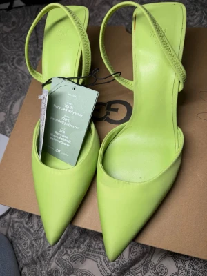 Limegröna pumps med rem från H&M - Snygga limegröna pumps med spetsig tå och smal rem runt hälen. Tillverkade i 100% återvunnen polyester med stilren design och klack. Perfekta för dig som vill sticka ut med färg och trendig look.