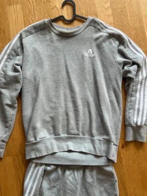 Grått Adidas loungewear set - Mysigt grått loungewear set från Adidas med klassiska vita ränder längs ärmar och ben. Setet består av en sweatshirt med rund hals och matchande byxor med resår i midjan. Adidas-logga på både tröja och byxa. Perfekt för chill dagar hemma.