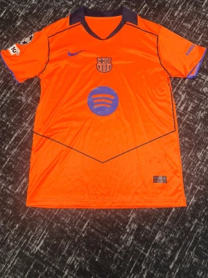 Orange FC Barcelona matchtröja - Snygg orange FC Barcelona matchtröja med lila detaljer och klubbmärke på bröstet. Tröjan har Spotify-logga på magen och Champions League-märke med siffran 5 på ärmen. Perfekt för fotbollsfans som vill sticka ut på planen eller läktaren.