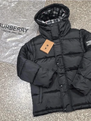 Svart pufferjacka från Burberry - Snygg svart pufferjacka från Burberry med huva och rutigt foder i grått, vitt och rött. Jackan har dragkedja och knappar framtill samt två sidofickor. Märkeslogga på ärmen och klassisk Burberry-känsla. Perfekt för kalla vinterdagar.