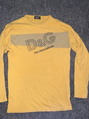 Gul långärmad D&G t-shirt - Storlek M-L Gul långärmad t-shirt från Dolce & Gabbana med stort D&G-tryck och diskreta ränder över bröstet. Klassisk rund halsringning och mjukt bomullsmaterial. Perfekt för dig som vill sticka ut med en ikonisk logga.