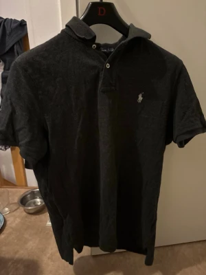 Svart pikétröja från Ralph Lauren - Klassisk svart pikétröja från Ralph Lauren med korta ärmar och den ikoniska loggan broderad på bröstet. Tröjan har krage och knappar framtill. Perfekt till jeans eller shorts för en clean look.