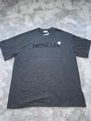 Svart Moncler t-shirt med logga - Snygg svart t-shirt från Moncler med stor ton-i-ton logga på bröstet och ett klassiskt Moncler-märke på vänster sida. T-shirten har rund hals och korta ärmar, tillverkad i mjuk bomull för en skön känsla.