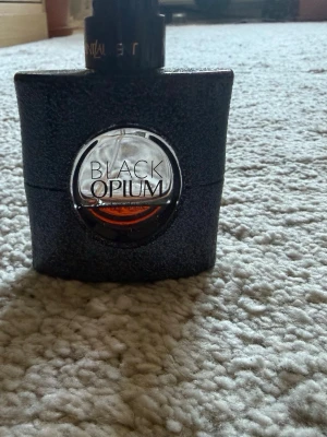 Black Opium parfymflaska 30 ml - Säljer en Black Opium parfym från Yves Saint Laurent. Flaskan är svart med glittrig yta och har ett runt fönster i mitten där den bärnstensfärgade vätskan syns. Modern och stilren design som känns lyxig och trendig.