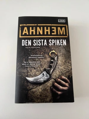 Den sista spiken av Stefan Ahnhem - Spännande kriminalroman med svart och gul omslag, skriven av Stefan Ahnhem. Boken är den sjätte delen om Fabian Risk och utgiven av Bokförlaget Forum. Omslaget pryds av en hand som håller i en kniv mot en mörk bakgrund.