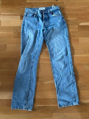 Ljusblå raka jeans med hög midja - Säljer ett par ljusblå jeans med rak passform och normal midja. Jeansen har klassiska fem fickor, bälteshällor och knappgylf. Materialet är denim i 100% bomull och de har en avslappnad vintage-look och har små slitningar. Finns inga bilder när jag bär då de inte passar långre!
