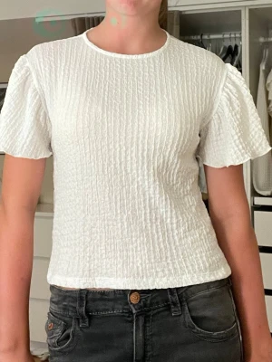 Vit strukturerad blus med kort ärm - Säljer en vit blus med strukturerat tyg och korta, lite puffiga ärmar.💕 Toppen har rund halsringning och en liten knappdetalj bak i nacken. Perfekt till jeans eller kjol för en fräsch look.Jag säljer den för att den är förliten och därför är den lite kort på mig på bilderna😅💕